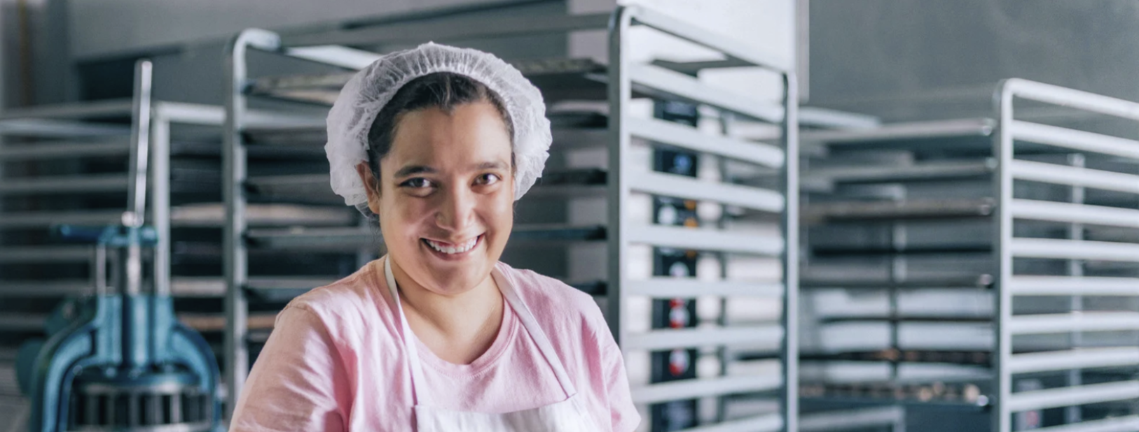 Smiling person in a bakery, wearing a hairnet and apron, stands in front of metal racks filled with trays.