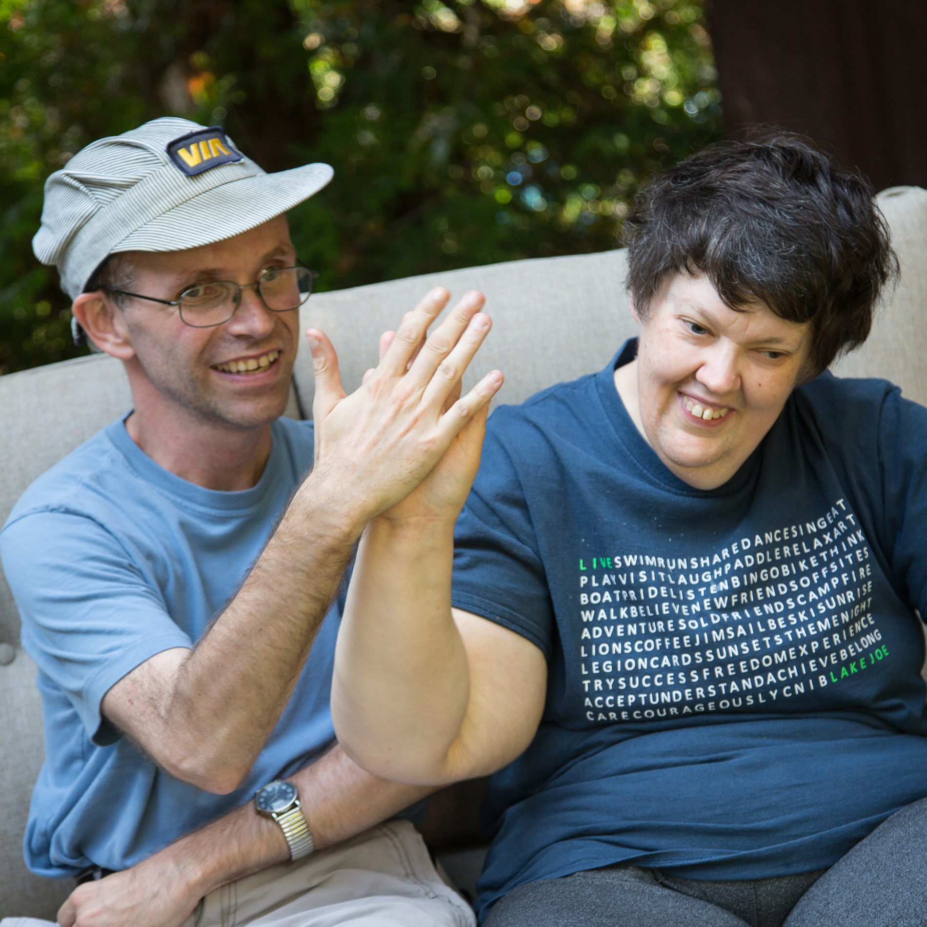 Two people smiling and high-fiving each other while sitting on a sofa outdoors. They appear joyful and relaxed in a casual setting.