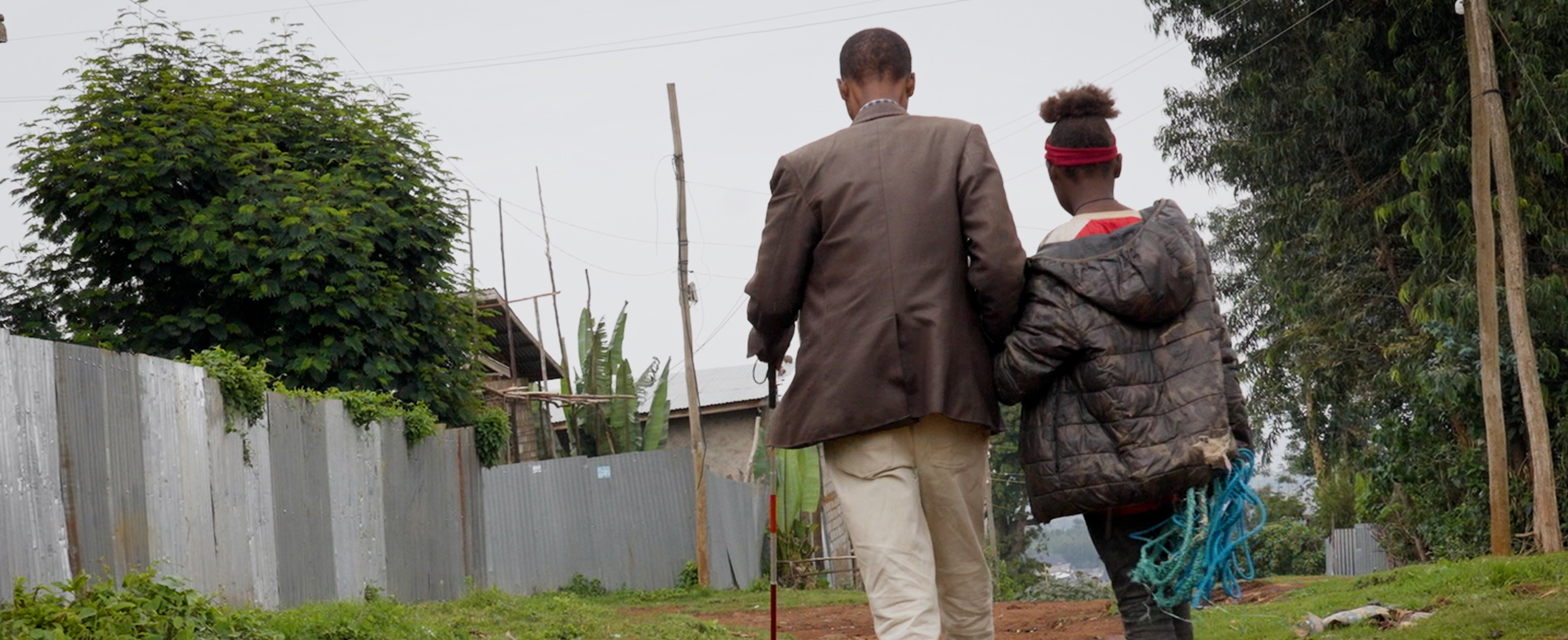 A man and a child walk down a dirt path lined with metal fences and trees, the man holds a cane and the child wears a dark jacket.