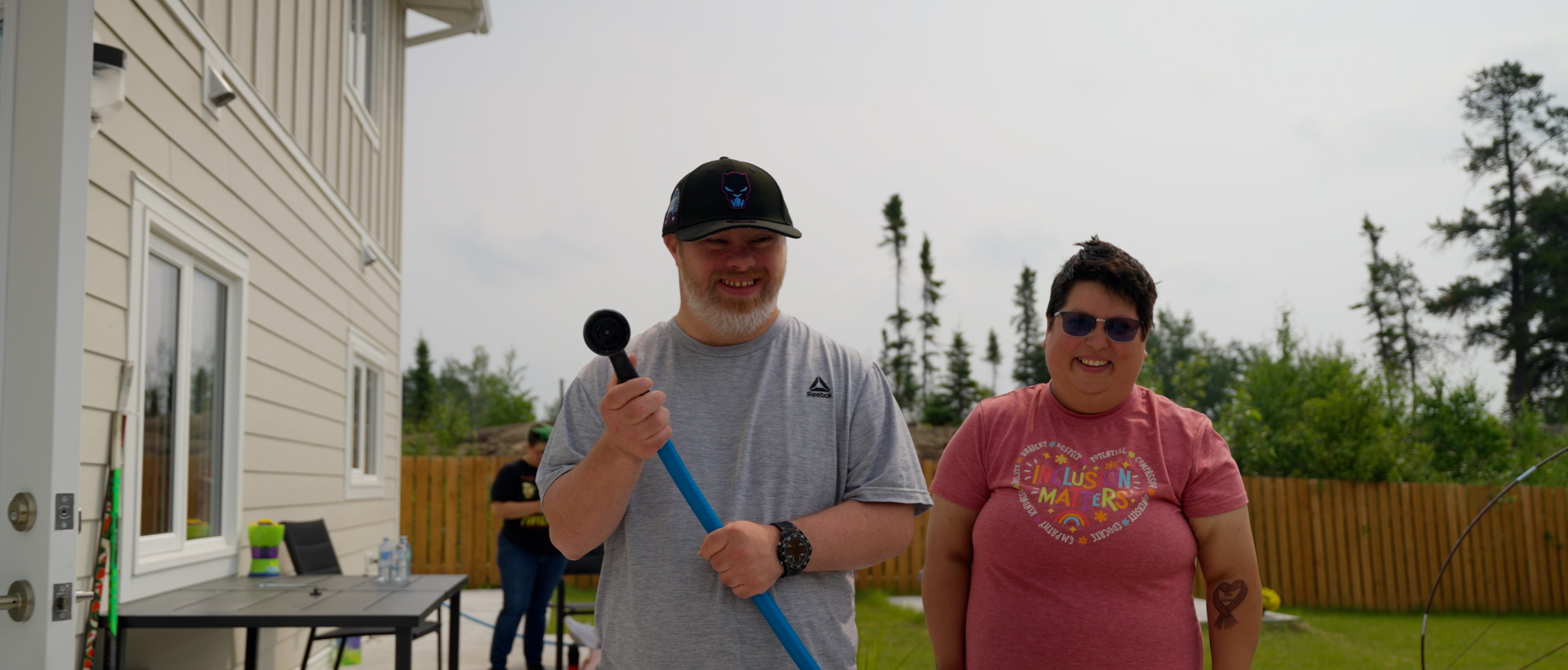 Two people smiling, one holding a hose in a backyard with trees. A table and chairs are visible by a light-colored house.