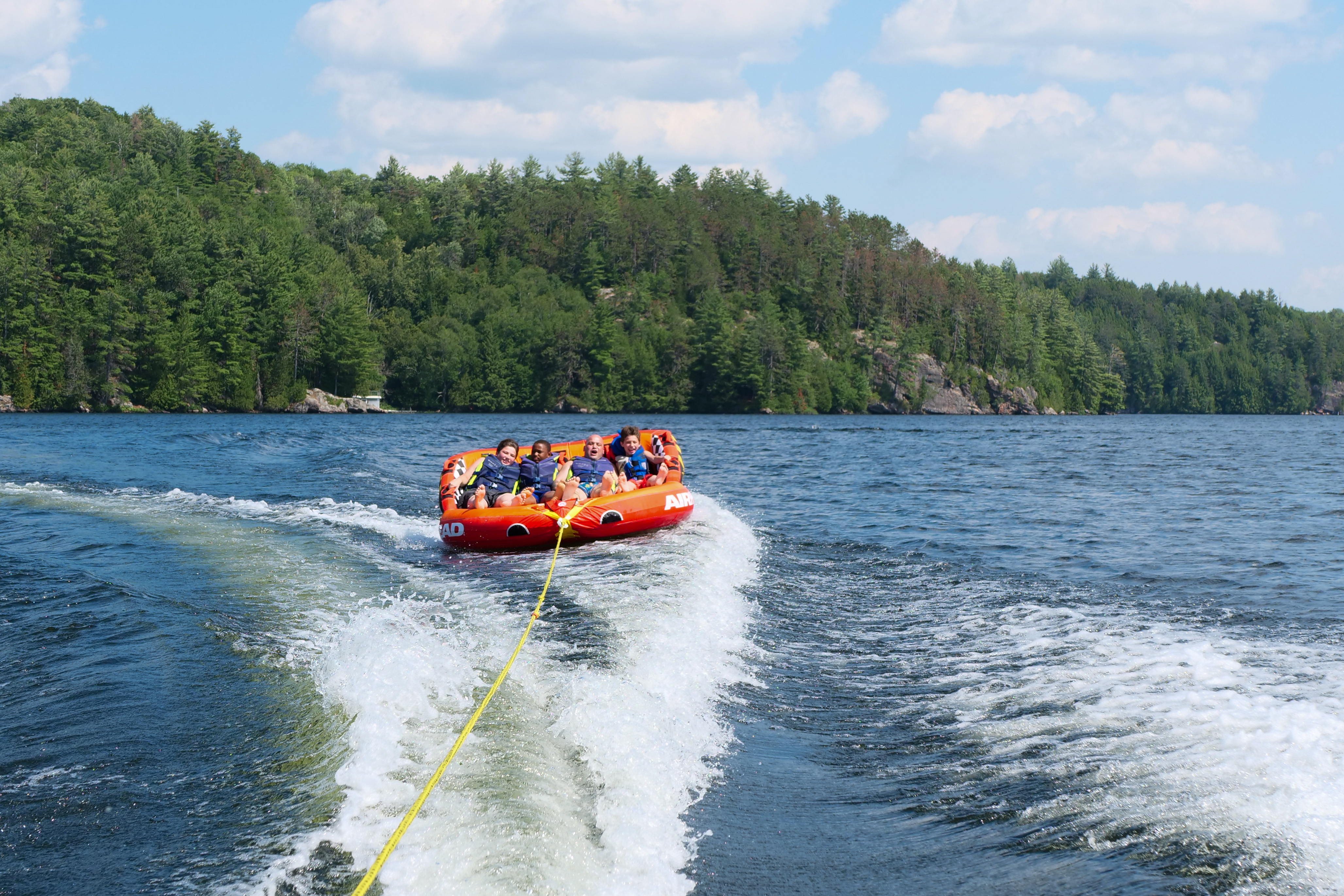 Four people ride an inflatable tube on a lake, pulled by a boat, with forested shoreline and a blue sky in the background.