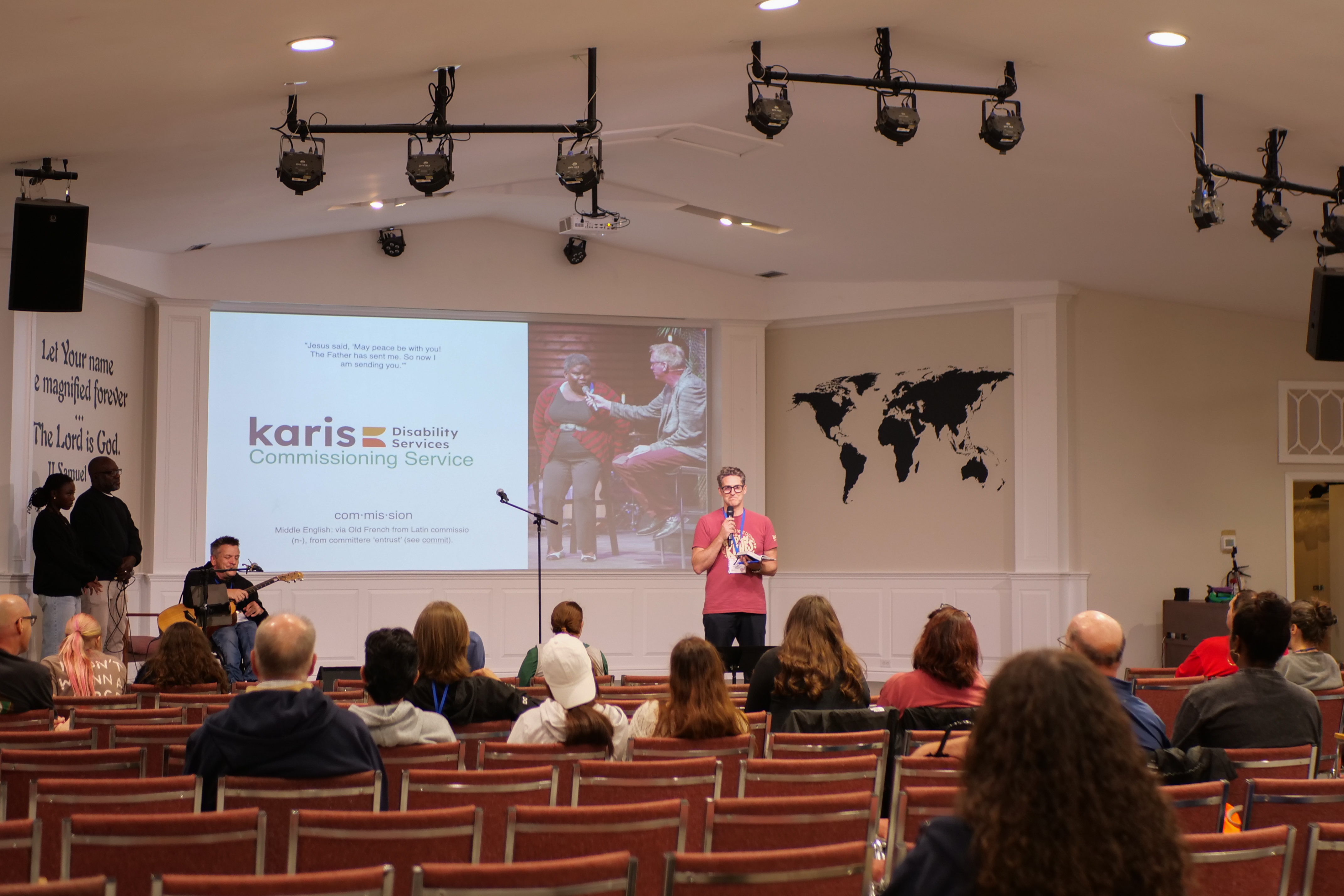 A speaker presents at the front of a church with a projected slide about "Karis Disability Services Commissioning Service." Audience seated in chairs.