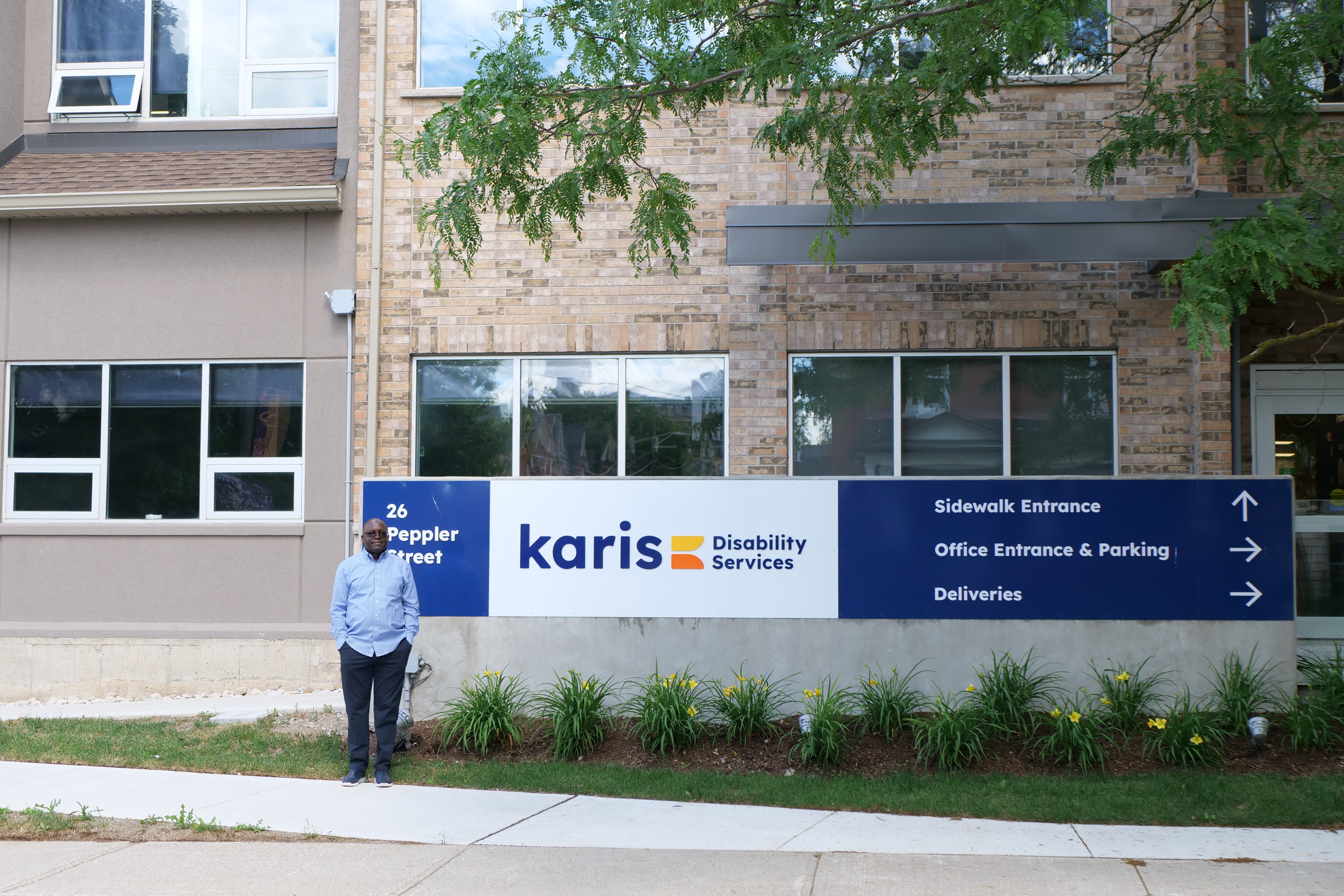 A person stands in front of a building with a "karis Disability Services" sign, surrounded by windows and greenery.