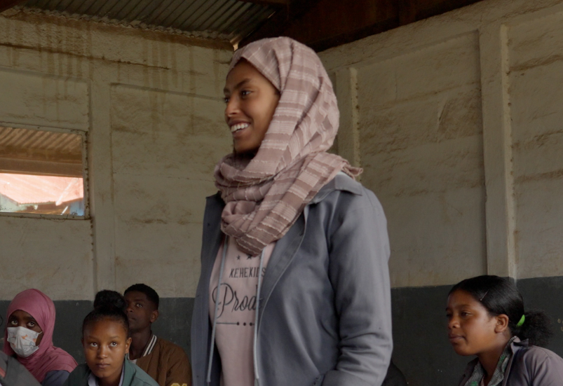 A woman in a classroom stands and smiles, wearing a patterned headscarf and gray jacket, surrounded by seated students taking notes.