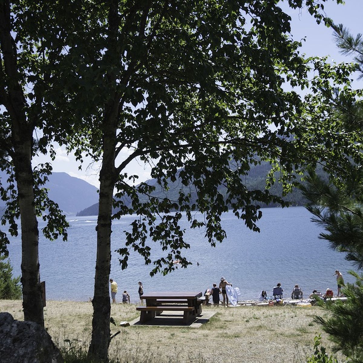 A serene lakeside scene with a picnic table under trees, people enjoying the beach, and mountains in the background.