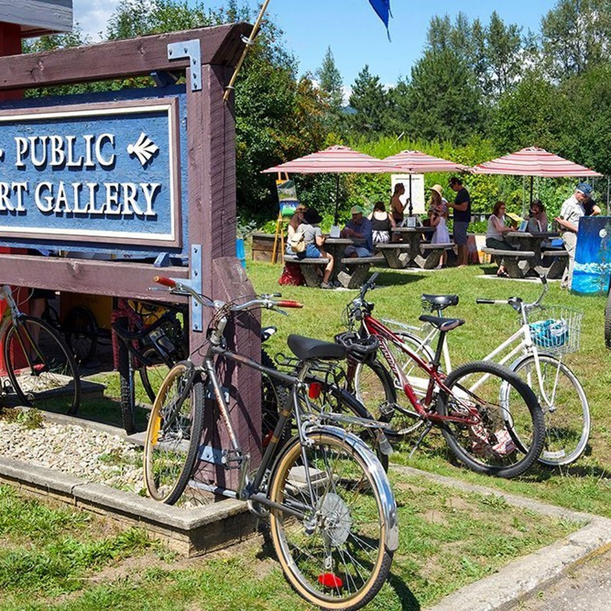 Bicycles parked by a "Public Art Gallery" sign, with people sitting at picnic tables under red-striped umbrellas in a grassy area.
