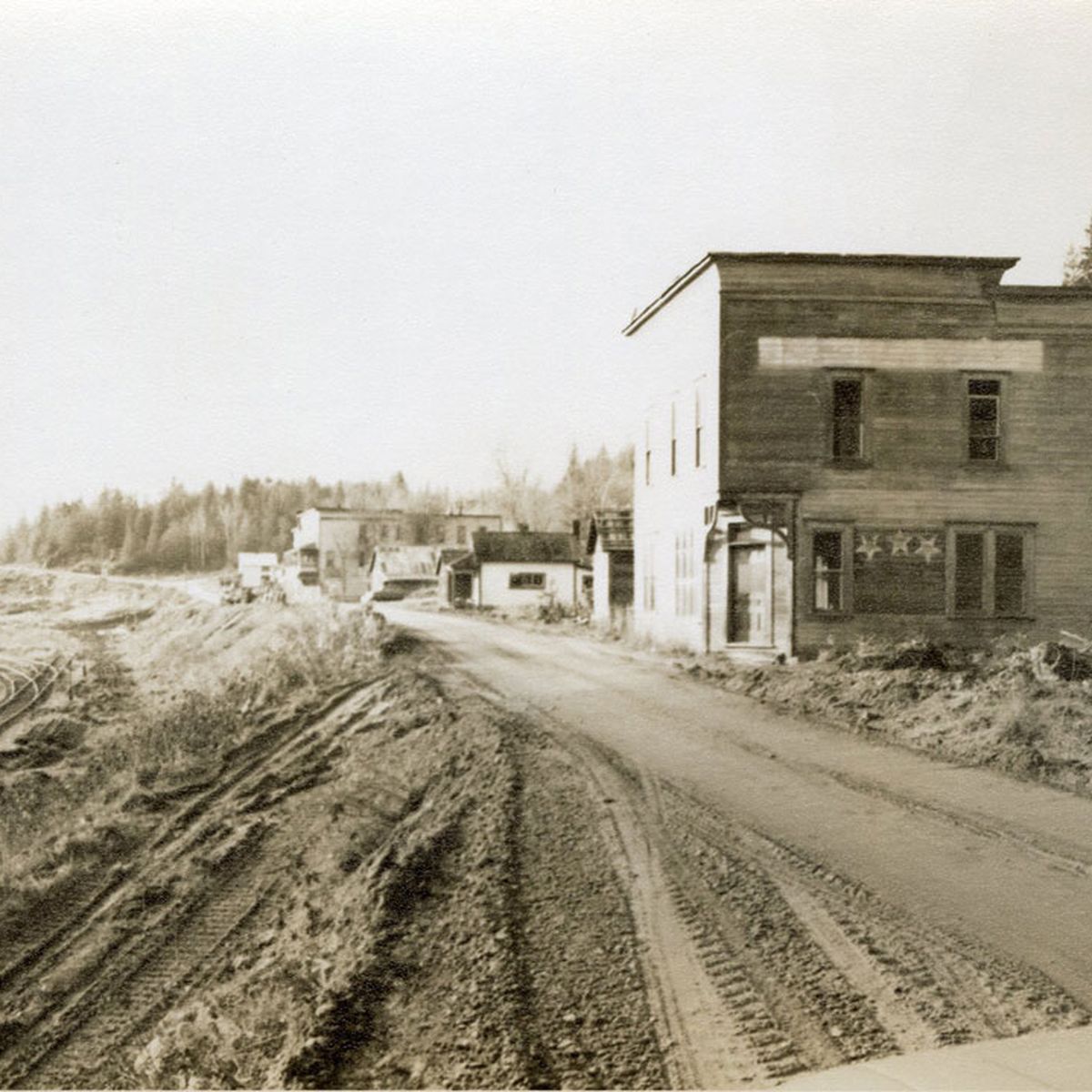 Sepia-toned image of a dirt road beside railroad tracks, with old wooden buildings and trees in the background.