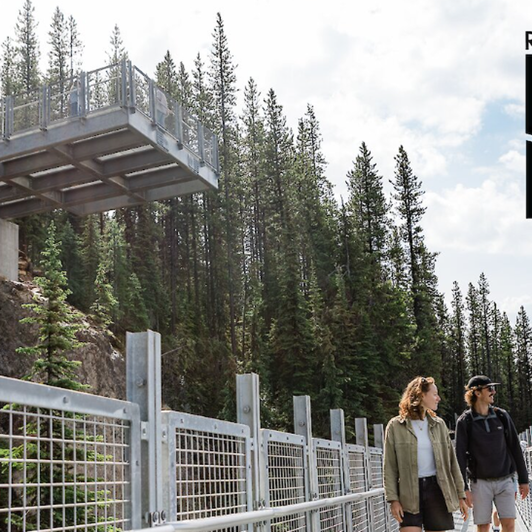 Two people walk on a metal walkway surrounded by pine trees, with a viewing platform above. Text reads "Revelstoke High Line."