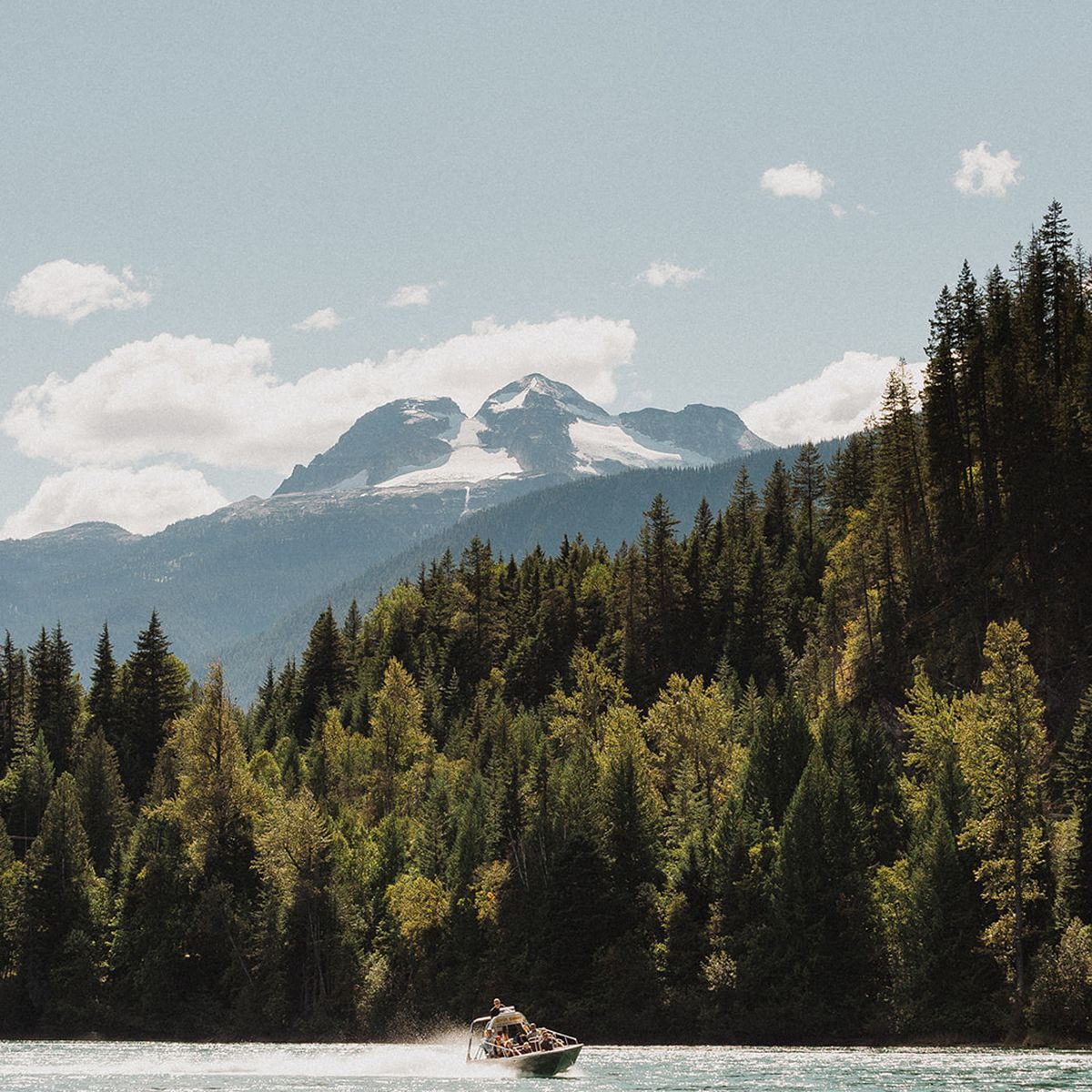 A boat speeds across a tranquil lake surrounded by dense forest, with majestic snow-capped mountains under a clear blue sky in the background.