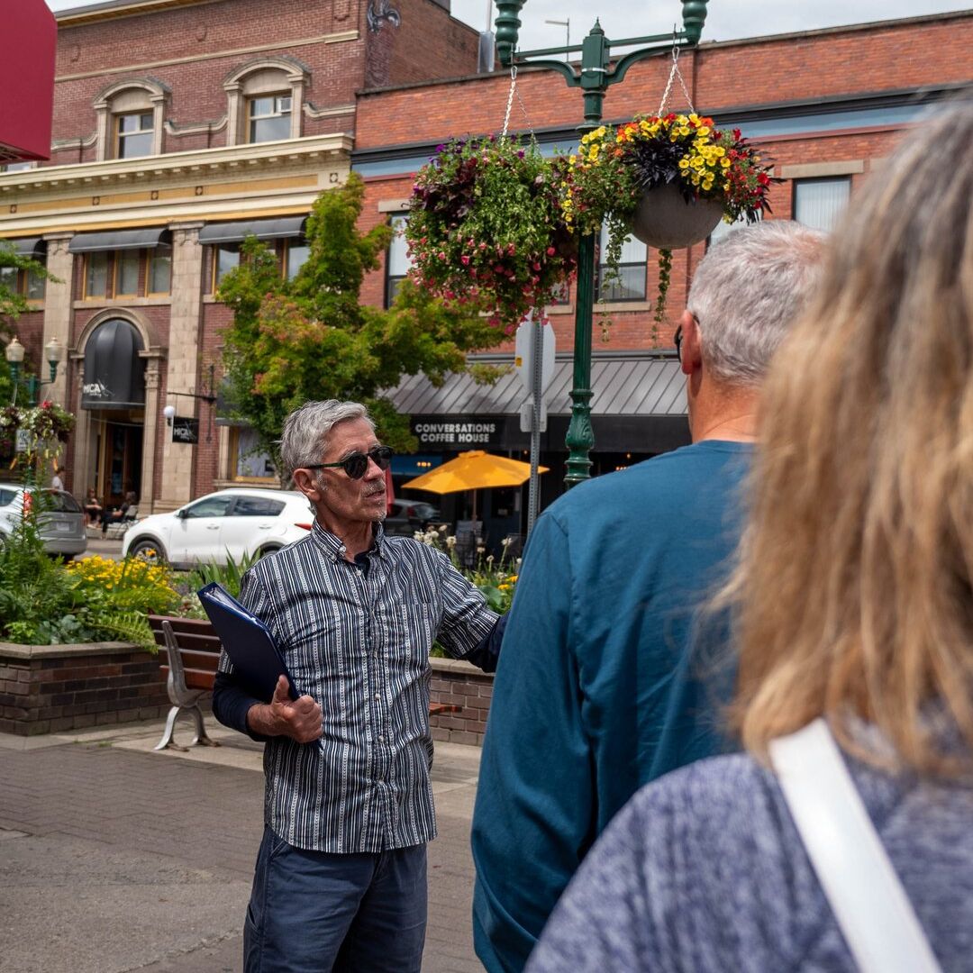 A man gives a tour to a small group on a city street, near a coffee house, with hanging flower baskets and historic buildings.