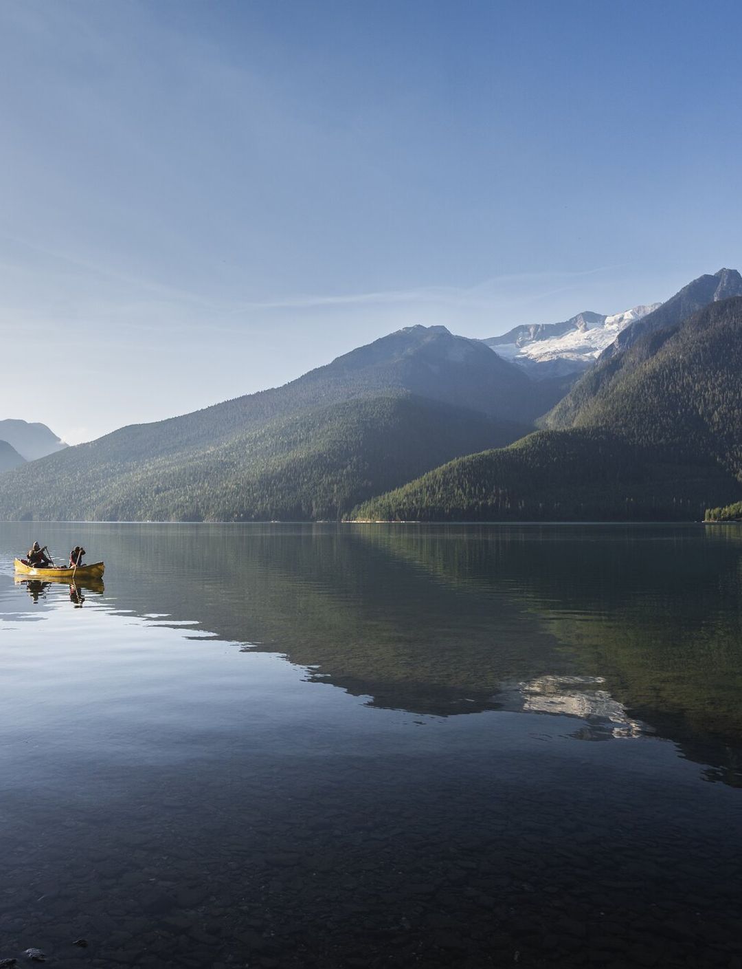 A yellow canoe with two people paddles on a calm lake, surrounded by forested mountains and a clear blue sky.