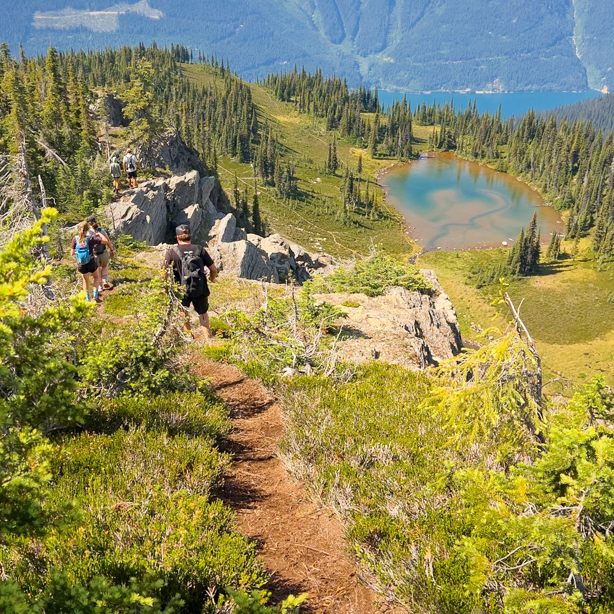 Hikers on a narrow trail overlooking a small lake and vast mountain range under a partly cloudy sky.