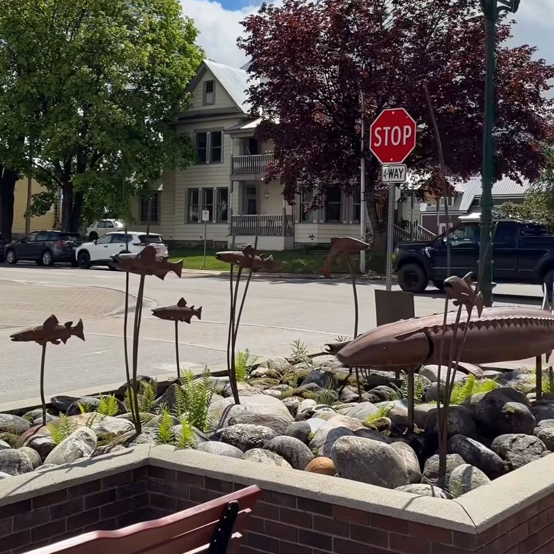 Metal fish sculptures in a landscaped rock garden near a street with a stop sign, trees, a house, and a lamp post in the background.