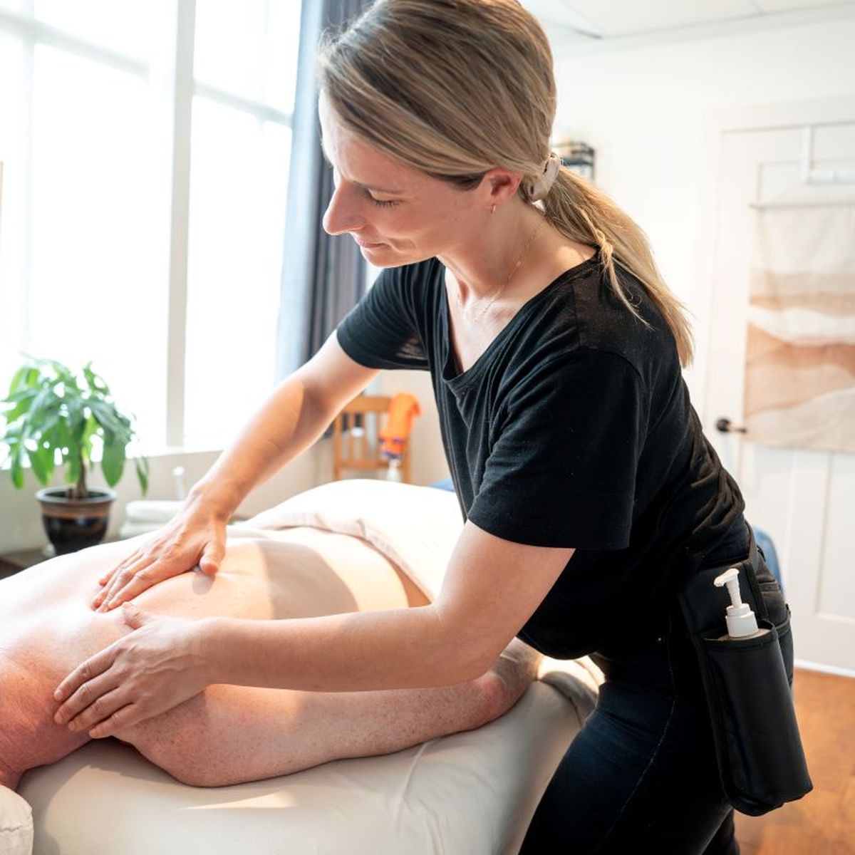 Person receiving a back massage in a bright room with a plant and a towel on the wall. The masseuse is wearing a black shirt.