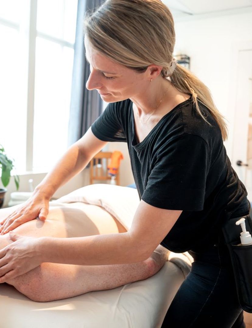 Person receiving a back massage in a bright room with a plant and a towel on the wall. The masseuse is wearing a black shirt.