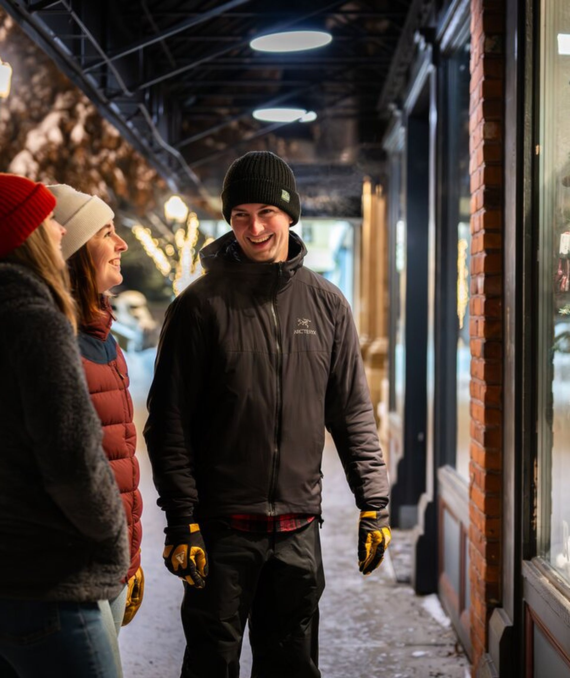 Three people in winter clothing smile while window shopping on a snowy, decorated street at night.