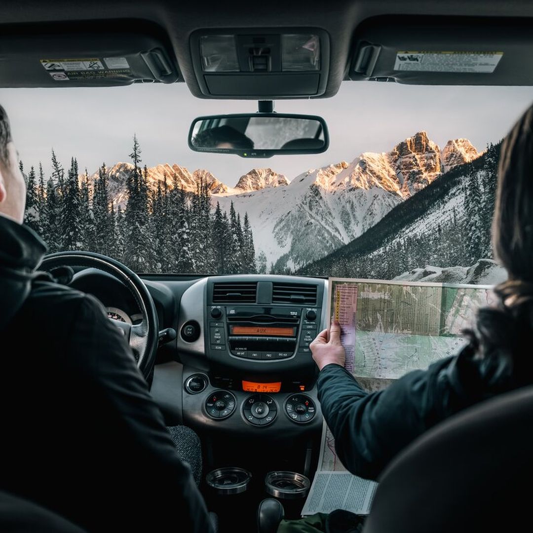 Two people in a car interior, with one holding a map, view snow-covered mountains through the windshield.