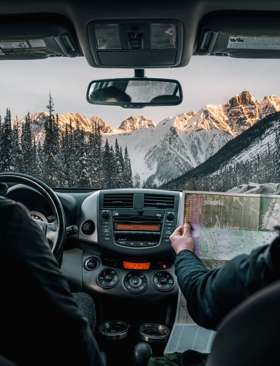 Two people in a car interior, with one holding a map, view snow-covered mountains through the windshield.