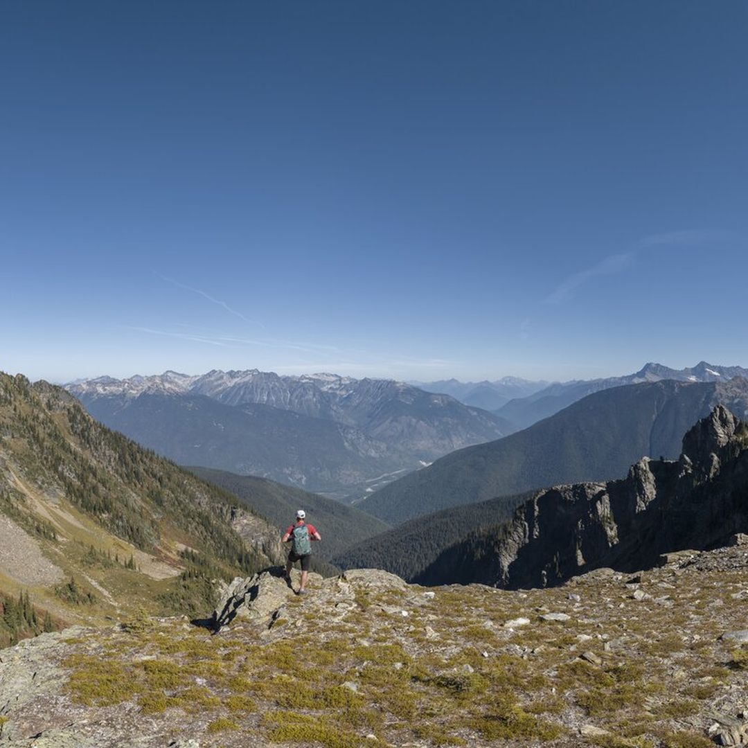 A hiker stands on a rocky mountain ridge, overlooking a vast valley with distant peaks under a clear blue sky.