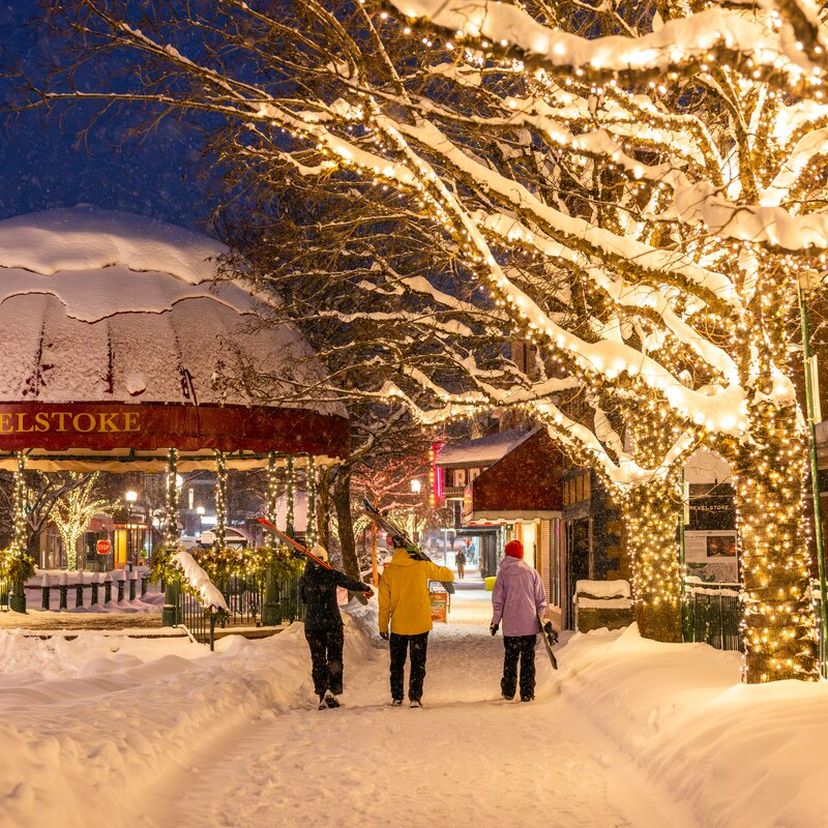 Snow-covered street with three people walking, surrounded by illuminated trees and a gazebo labeled "Revelstoke" at night.
