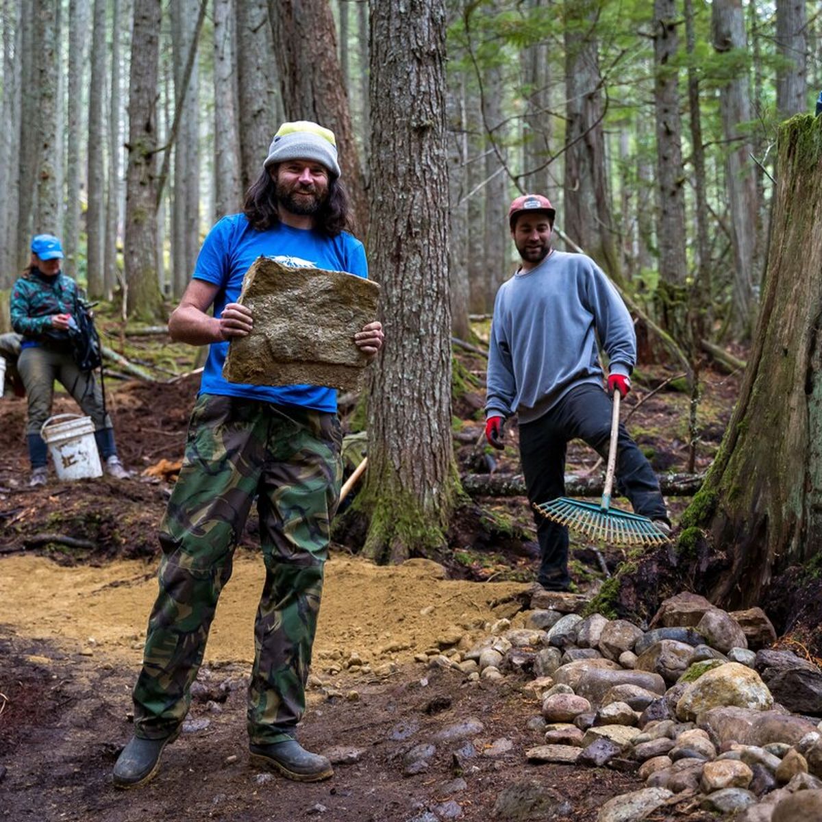 Three people working on a forest trail; one holds a rock, another holds a rake, surrounded by trees and dirt.