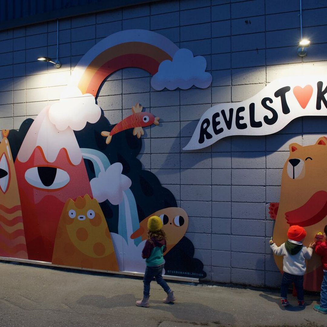 Colorful mural with playful mountain, rainbow, and bear designs. Two children engage with the artwork under bright spotlights.