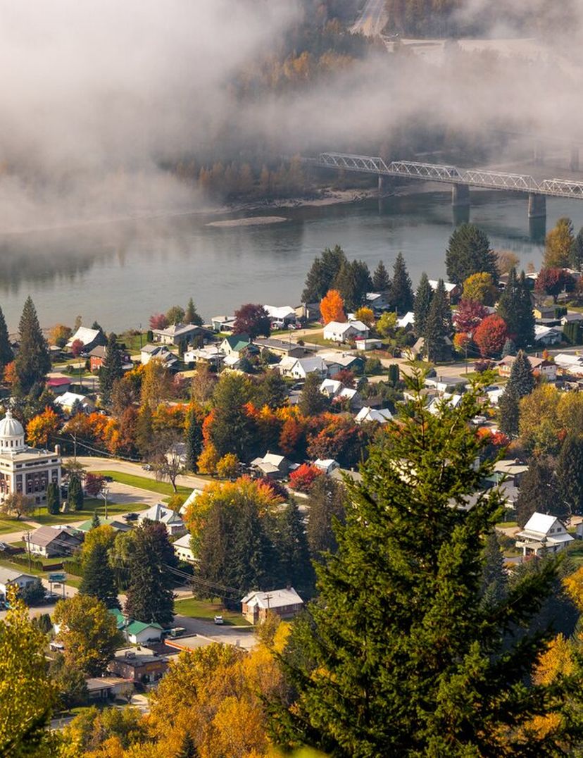 Aerial view of a town with colorful autumn trees, a river with a bridge, and misty hills in the background.