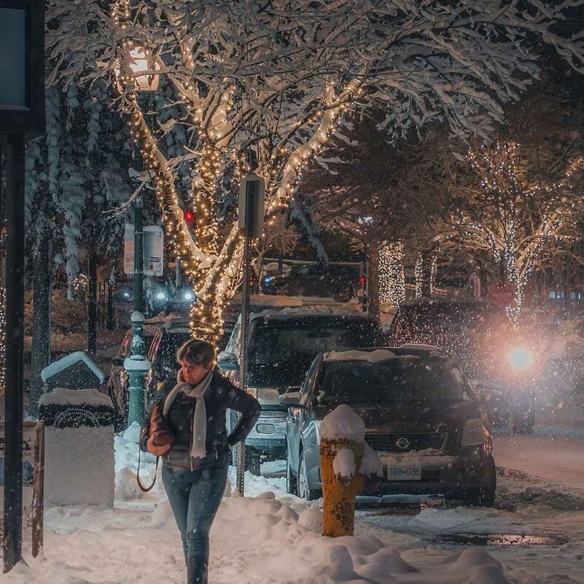 Person walking on a snowy street at night, with trees wrapped in string lights and cars parked along the road.
