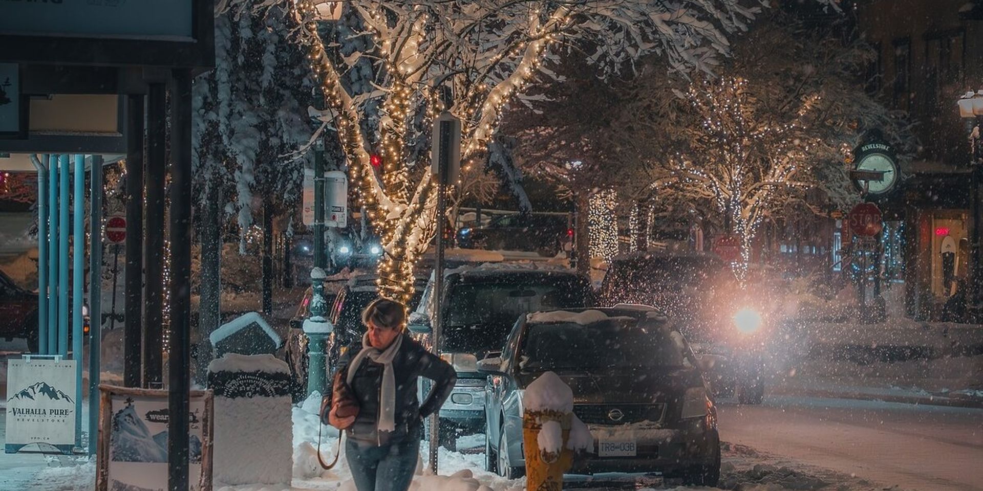Person walking on a snowy street at night, with trees wrapped in string lights and cars parked along the road.