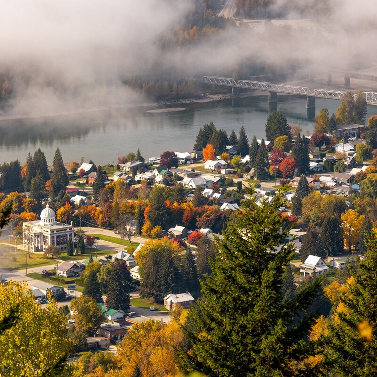 Aerial view of a small town with colorful autumn trees, a river, and a bridge, surrounded by misty hills and forested areas.