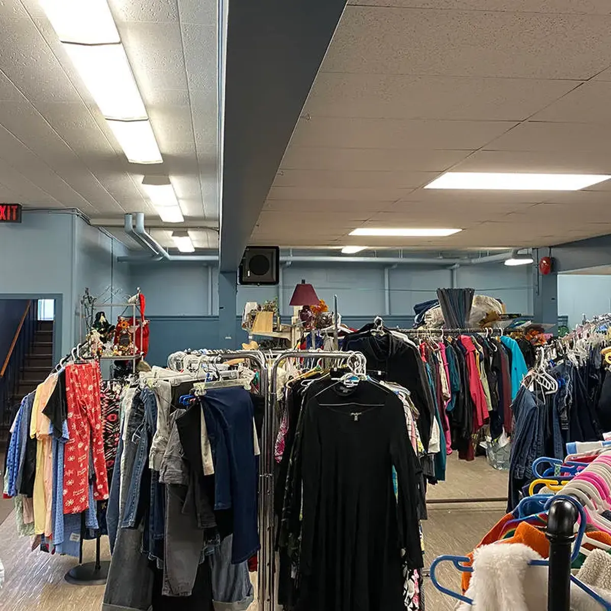 Thrift store interior with racks of assorted clothing and items, under fluorescent lights and blue walls, with an exit sign visible.