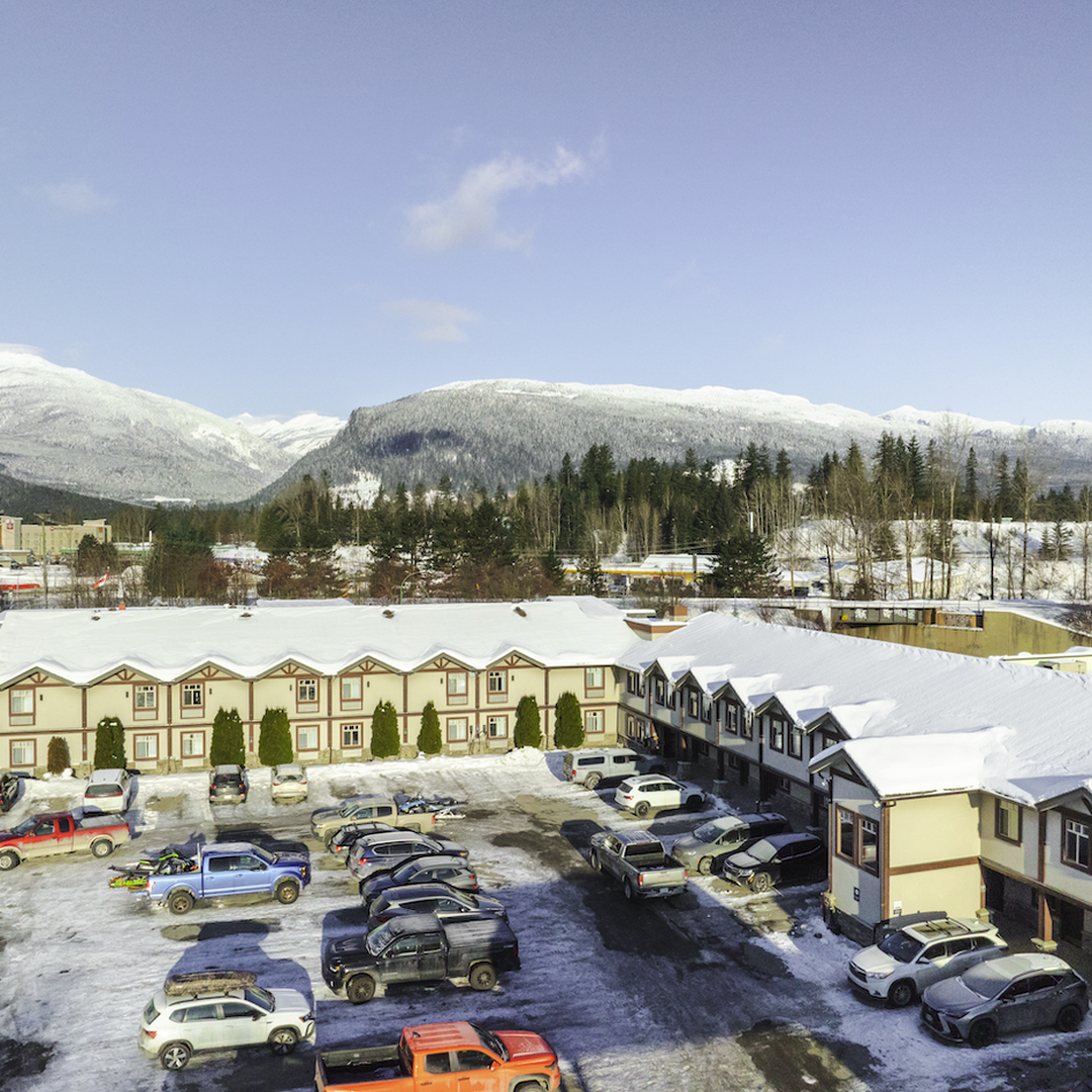 Snow-covered buildings and parked cars in a snowy landscape, with forested mountains in the background under a clear blue sky.