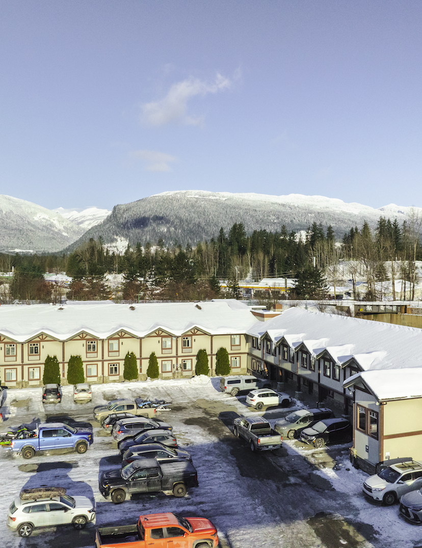 Snow-covered buildings and parked cars in a snowy landscape, with forested mountains in the background under a clear blue sky.