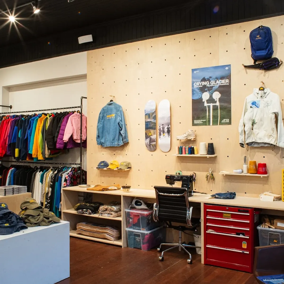 Clothing store interior with colorful jackets on racks, hats on a table, a workspace with a chair, and wall decor including posters and skateboards.