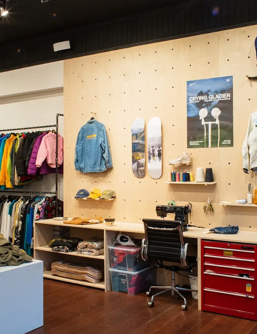 Clothing store interior with colorful jackets on racks, hats on a table, a workspace with a chair, and wall decor including posters and skateboards.