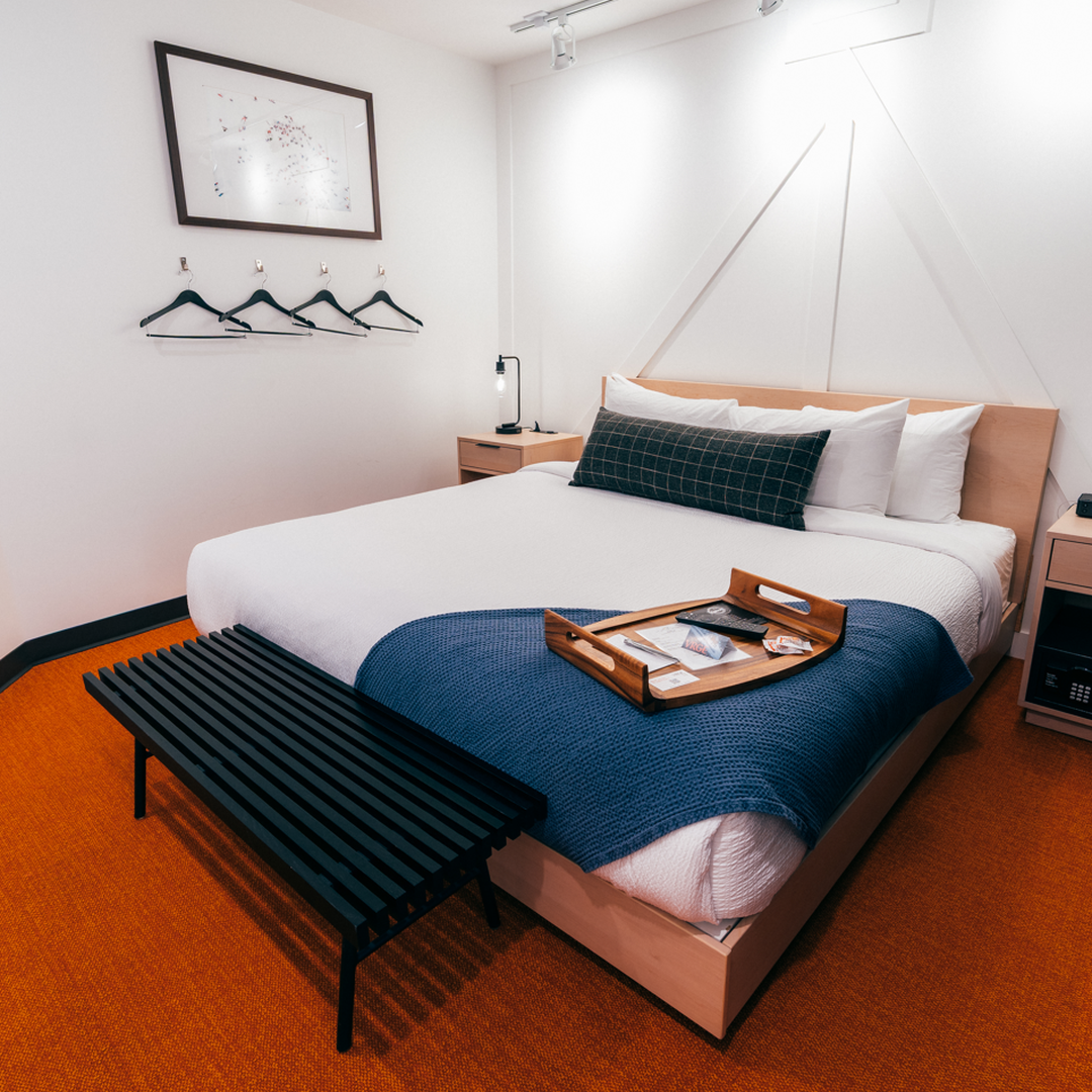 Modern hotel room with a neatly made bed, blue blanket, and tray. Nightstands with lamps, wall hooks, and a painting decorate the white walls.