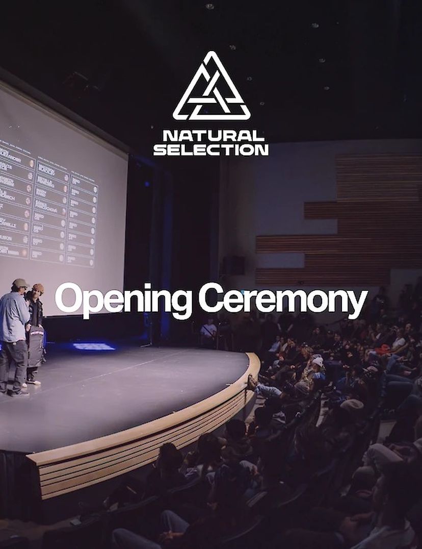 Wide-angle view of a stage with two speakers, large screen, and audience. "Natural Selection Opening Ceremony" is projected on the screen.