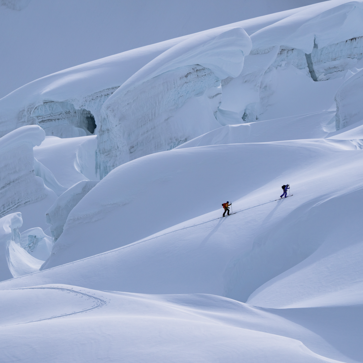Two climbers ascend a snowy mountain slope amidst large ice formations under a clear blue sky.