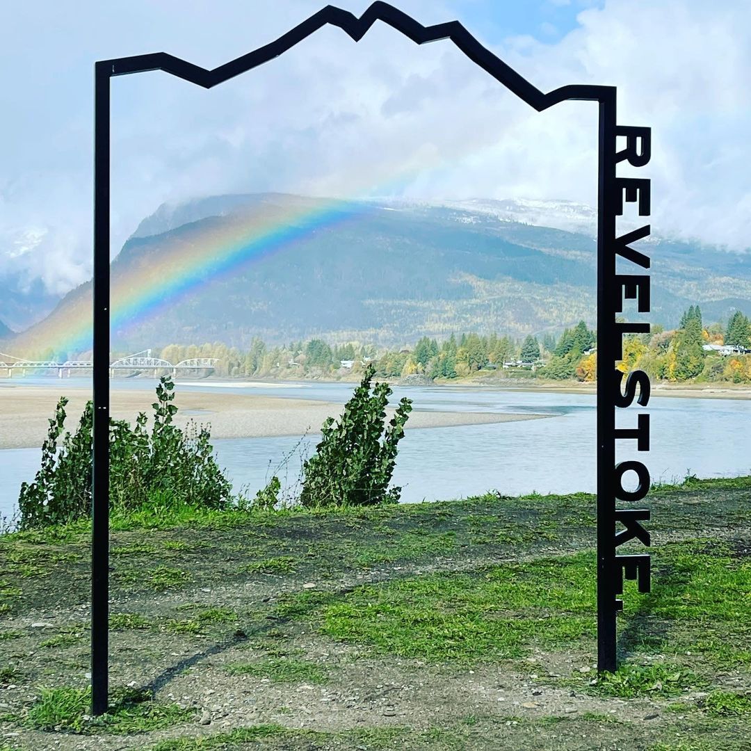 Black frame with "Revelstoke" text facing a vibrant rainbow over a river, with mountains and trees in the background under a cloudy sky.