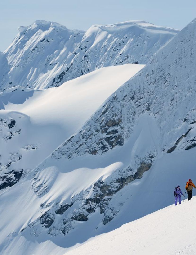 Two people hiking up a snowy mountain slope with jagged peaks in the background under a clear blue sky.