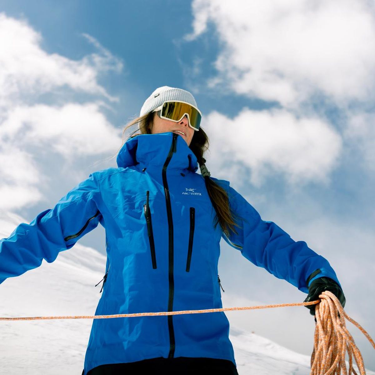 Person in blue jacket and goggles holding a climbing rope on a snowy mountain under a blue sky with clouds.