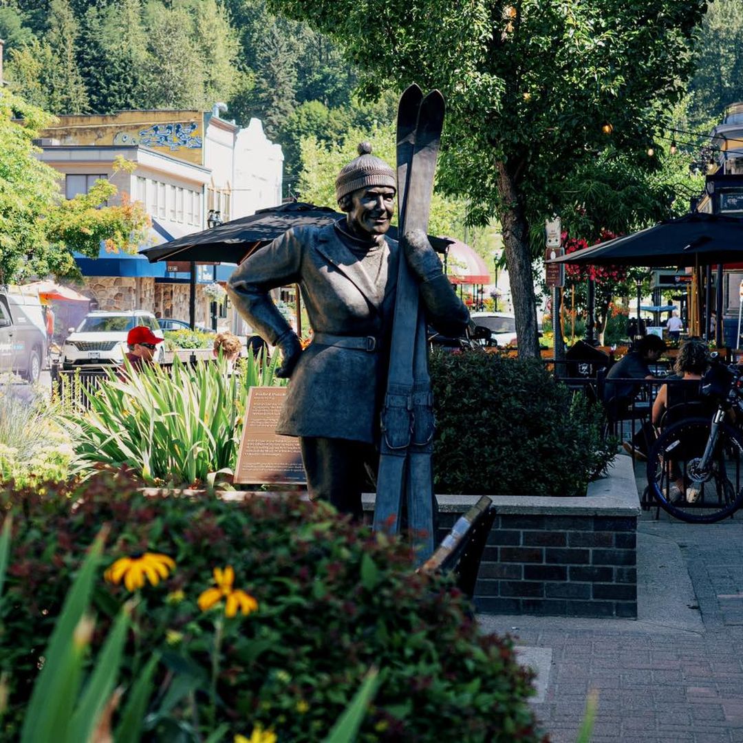 Statue of a skier with skis in a vibrant downtown area, surrounded by flowers, people dining, and parked cars.