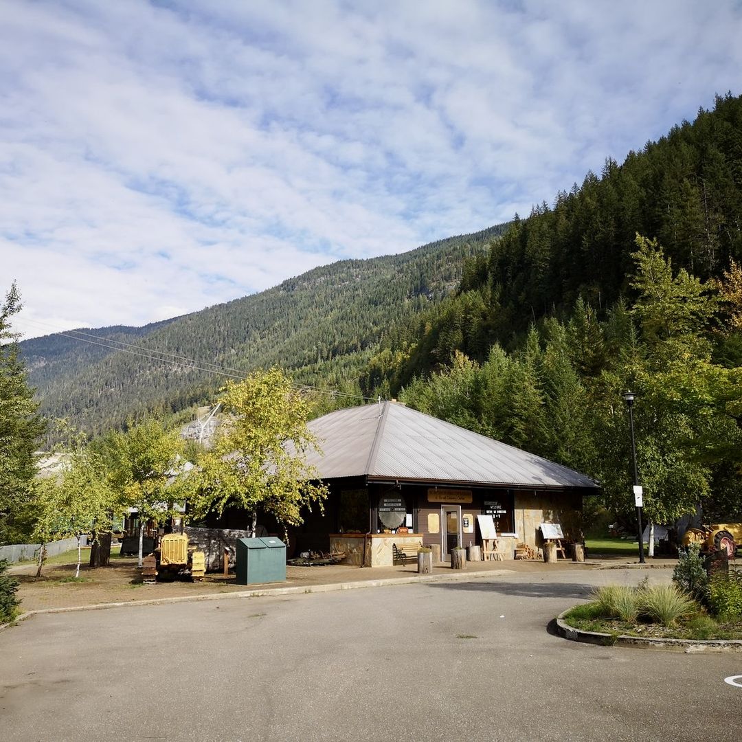 A rustic building with a sloped roof sits among trees and mountains under a partly cloudy sky, with a paved area in the foreground.