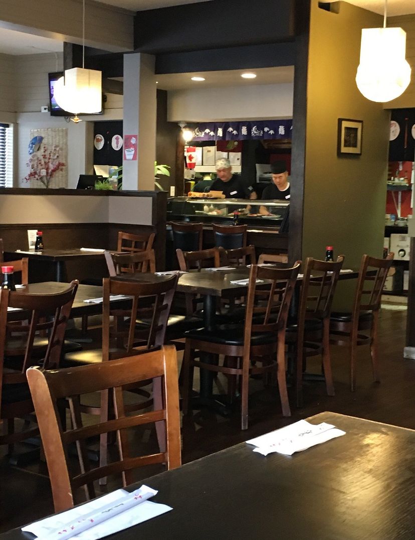 Cozy restaurant interior with wooden tables and chairs, sushi chefs working behind the counter, and soft lighting creating a warm ambiance.