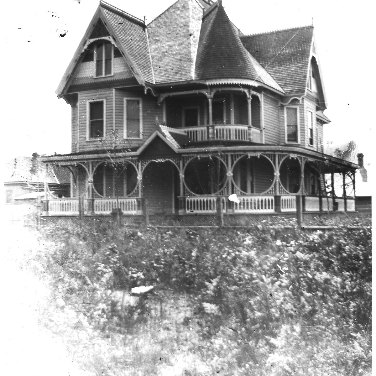 A vintage black-and-white photo of a large Victorian house with ornate wooden details, a wraparound porch, and steep gabled roofs.