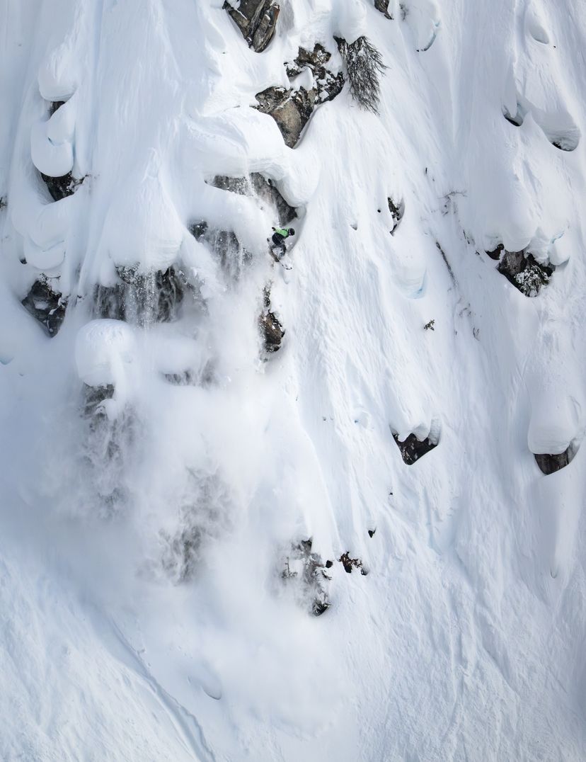 A skier makes their way down a steep, snow-covered mountain slope, surrounded by rocks and fresh powder snow.