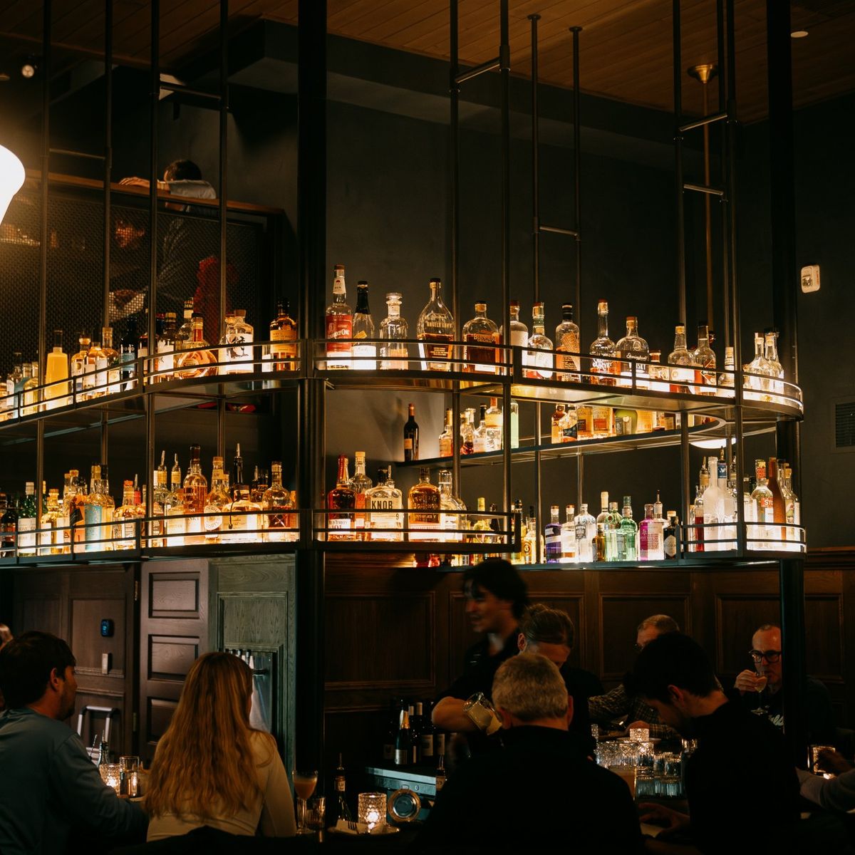Dimly lit bar with patrons seated around a counter. Shelves above display various bottles of liquor, illuminated by warm lighting.
