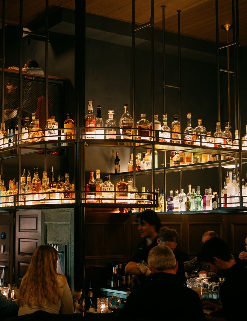 Dimly lit bar with patrons seated around a counter. Shelves above display various bottles of liquor, illuminated by warm lighting.