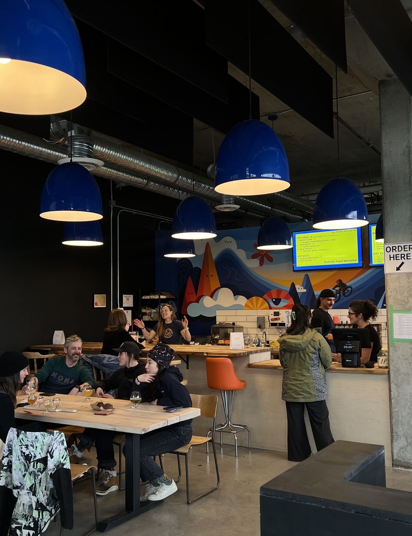 Modern brewery with blue pendant lights, colorful mural, and patrons enjoying drinks. Staff serve orders at the counter. Cozy and vibrant atmosphere.
