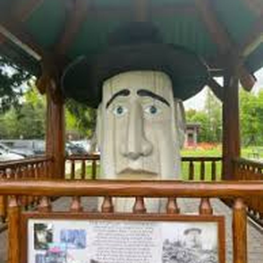 Large white face sculpture with a black hat under a gazebo, surrounded by wooden railings, with an informational plaque in front.